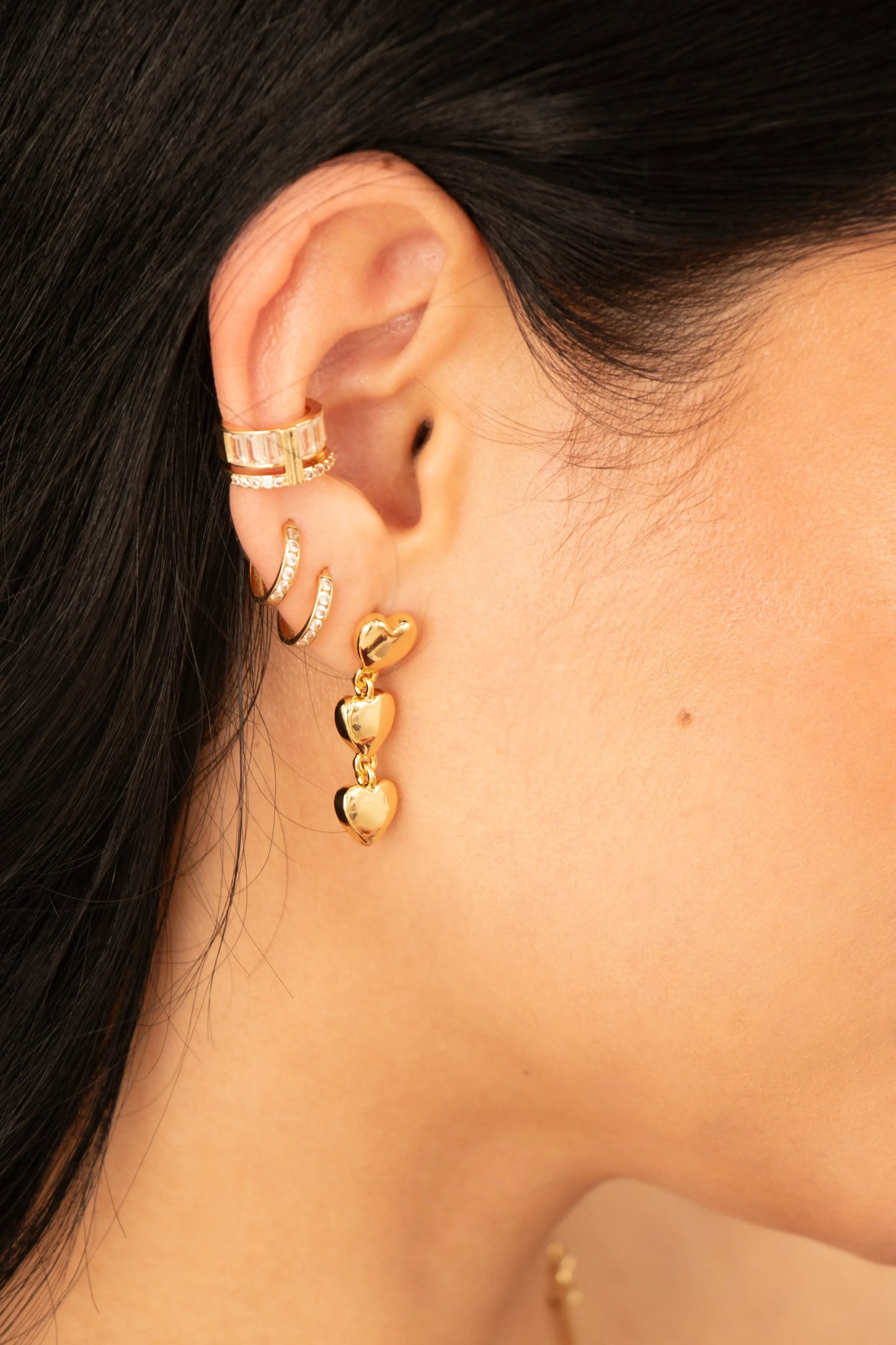 Aretes dorados colgantes de triple corazón en baño de oro 24K, fotografía en oreja de modelo.