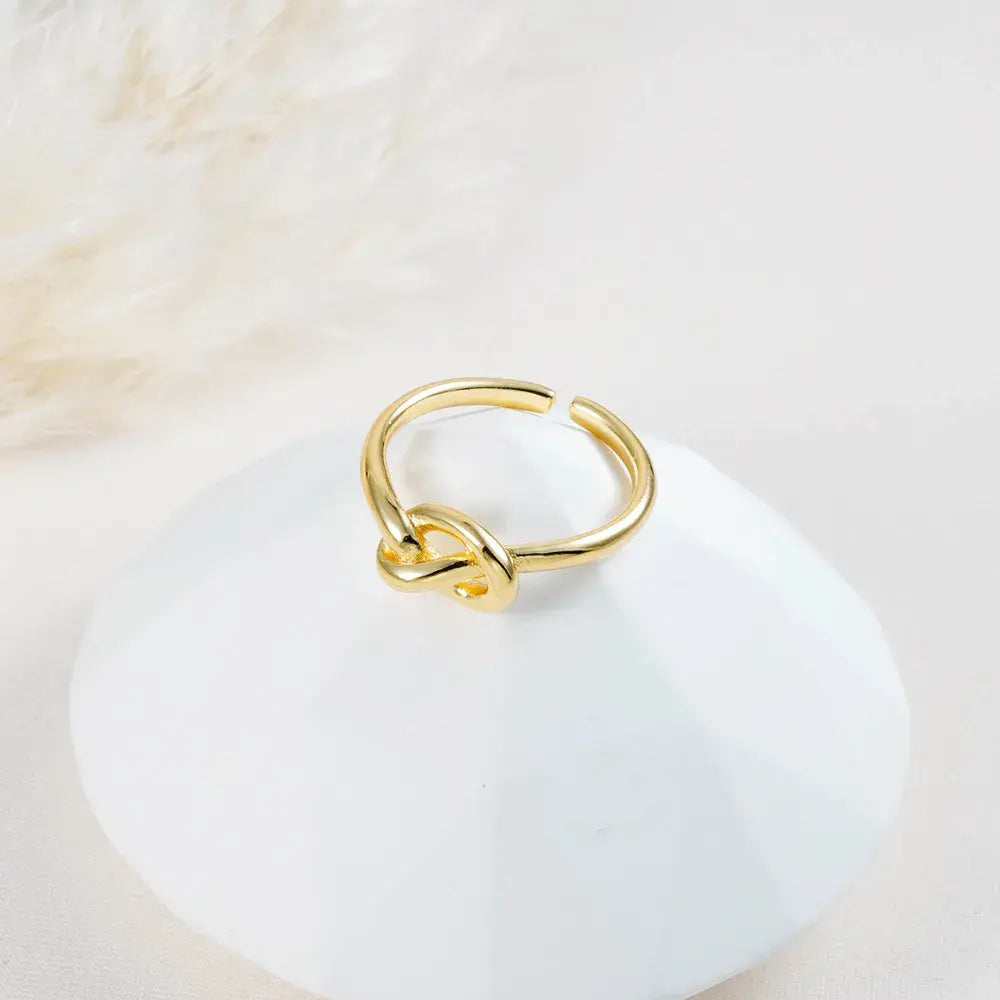 Anillo dorado ajustable con nudo central en baño de oro 24K, fotografía sobre base blanca.