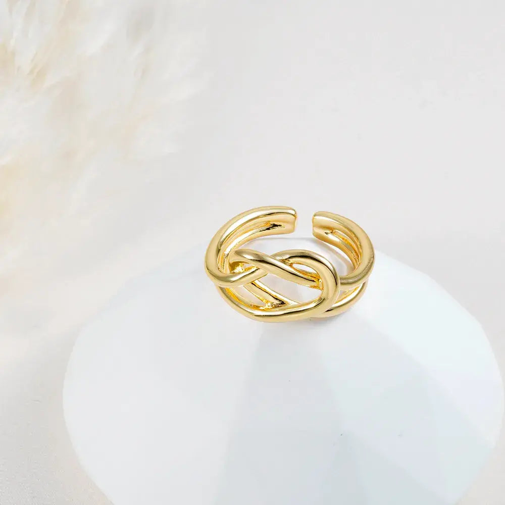 Anillo dorado ajustable con nudo entrelazado en baño de oro 24K, fotografía sobre base blanca.
