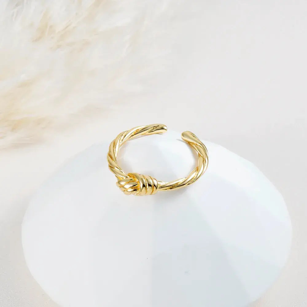Anillo dorado ajustable trenzado con nudo central en baño de oro 24K, fotografía sobre base blanca.