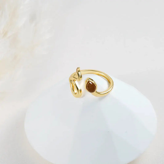 Anillo dorado tipo abierto con gota de color café en baño de oro 24K, fotografía sobre base blanca.