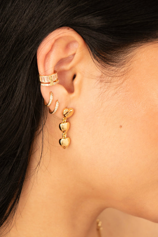 Aretes dorados colgantes de triple corazón en baño de oro 24K, fotografía en oreja de modelo.