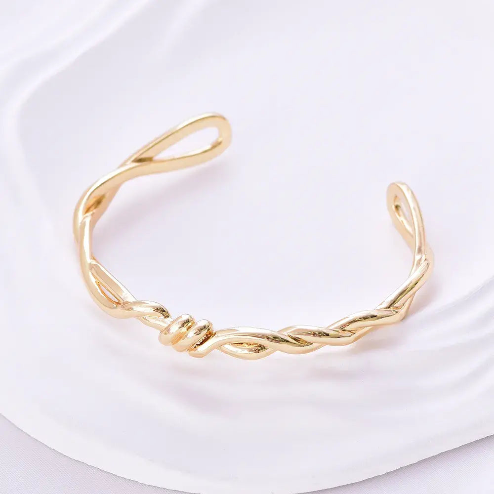 Brazalete rígido dorado con trenzado doble y nudo central, acabado brillante en baño de oro 24K.