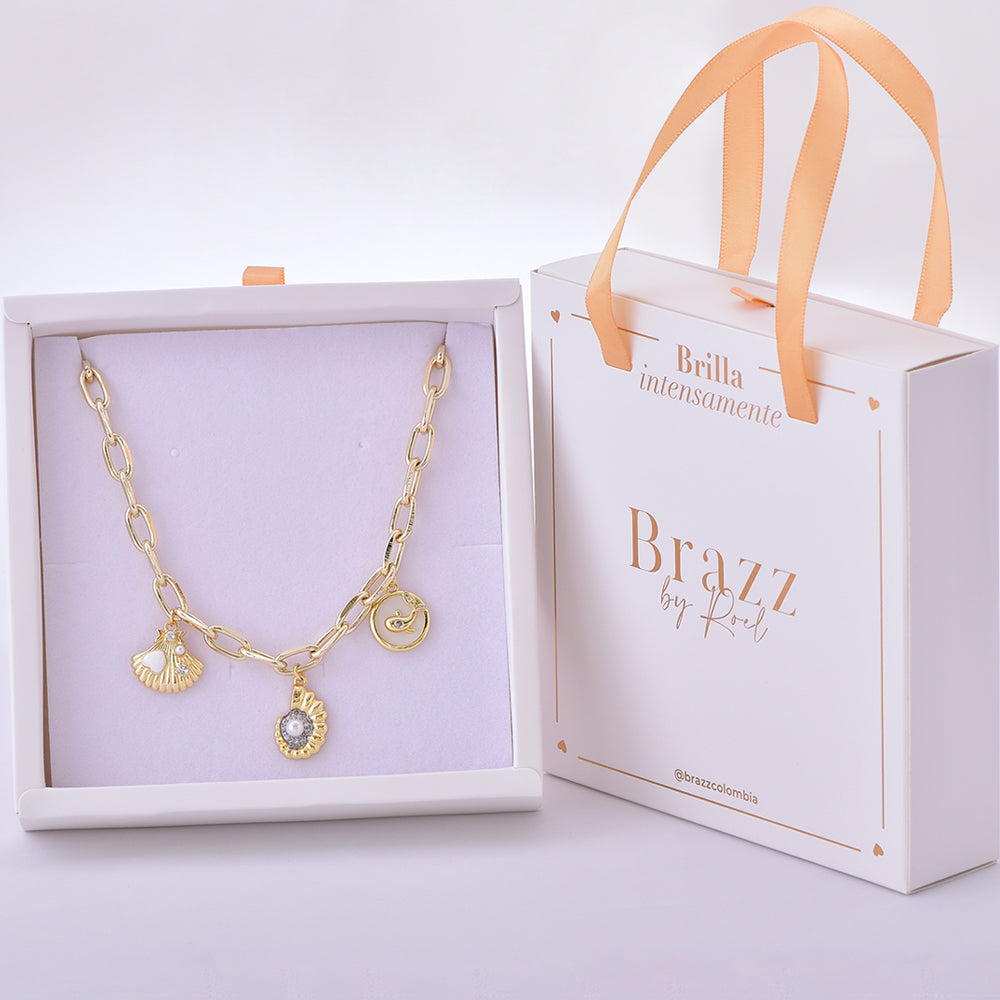 Collar dorado con dijes marinos, incluyendo concha y charms decorativos, en caja Brazz by Roel.