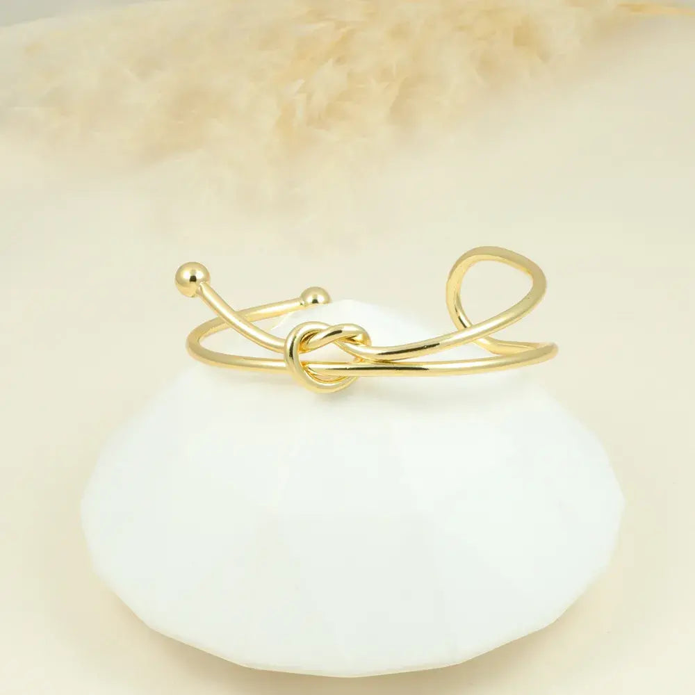 Pulsera abierta dorada con diseño de nudo sobre base blanca.